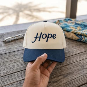 May include: A two-tone baseball cap featuring a cream-colored crown and a dark blue brim. The word Hope is embroidered in dark blue cursive script across the front center of the crown. The cap has a structured five-panel design with a top button.