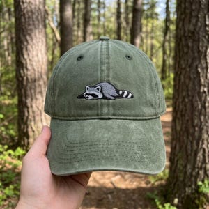 May include: An olive green baseball cap with a washed, distressed finish. The front panel features an embroidered patch of a grey and white raccoon lying down in a relaxed position, complete with its signature black eye mask and striped tail.