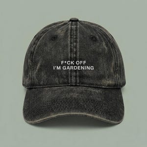 May include: A black acid-wash baseball cap with a curved brim and a button on top. The front of the cap features white embroidered text that reads F*CK OFF I'M GARDENING in a clean, sans-serif font. The cap has a six-panel construction.