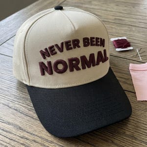 May include: A beige and black baseball cap with the words "NEVER BEEN NORMAL" embroidered in maroon. The cap has a black brim and a black button on top. A spool of maroon thread, a needle, and a pink fabric scrap are next to the cap.