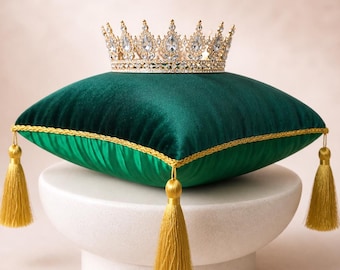 May include: A gold-toned metal crown featuring clear, teardrop-shaped crystals resting on a square, dark green velvet pillow. The pillow is trimmed with gold cord piping and features a gold tassel at each of its four corners for a decorative finish.