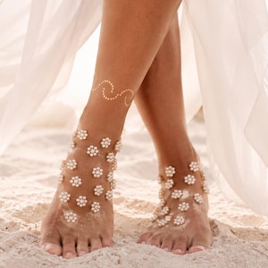 May include: A pair of barefoot sandals featuring white floral embellishments with gold centres, arranged in a delicate pattern across the top of the feet. A gold wave-patterned temporary tattoo is visible on the lower leg above the ankle.
