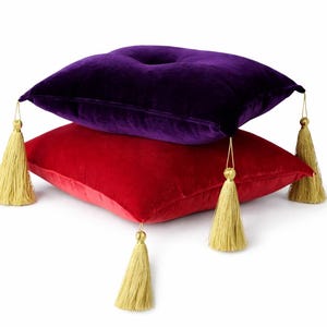 Puede incluir: Dos cojines de terciopelo, uno rojo y otro morado, apilados uno encima del otro. Cada cojín tiene una borla dorada en cada esquina. Los cojines están sobre un fondo blanco.