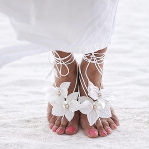 May include: White barefoot sandals with floral details and string ties. The sandals have large white flower embellishments with pearl centres. The feet are painted with pink nail polish, and the background is white sand.