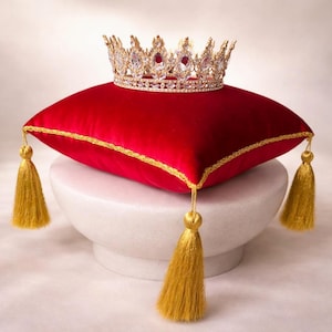 May include: A gold-toned metal crown featuring clear, teardrop-shaped crystals and small pavé stones, resting atop a square, deep red velvet cushion. The cushion is trimmed with gold piping and features a gold tassel at each of its four corners.