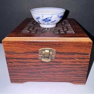 May include: A small white ceramic bowl with blue painted bamboo patterns sits atop a rectangular wooden box. The box features a dark brown wood grain finish, a metal latch on the front, and a decorative openwork lattice design on the top lid.