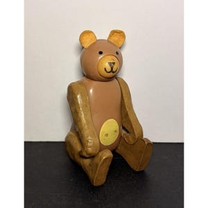 May include: A vintage brown and tan wooden bear toy with movable arms and legs. The bear has a round head with small ears, a black nose, and a yellow circle on its belly. The bear is sitting down.