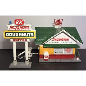 May include: A miniature Krispy Kreme shop and sign. The sign reads "DOUGHNUTS" and "COFFEE" with the "KK" logo. The shop has a green roof, yellow and red walls, and the text "Krispy Kreme DOUGHNUTS & COFFEE". A small tree is next to the shop.