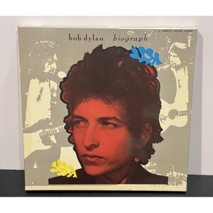 May include: A Bob Dylan "Biograph" cassette tape, deluxe edition. The album cover features a red and black portrait of Bob Dylan, with the album title in white text. The background is a grey and white design with images of Bob Dylan.