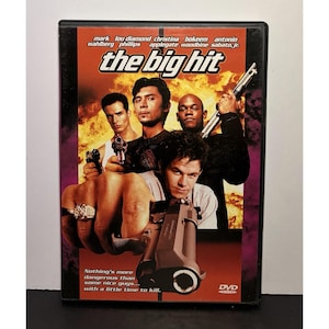 Op de afbeelding: DVD-hoes voor de film "The Big Hit" met een groep mannen met wapens tegen een vurige achtergrond. De titel staat in grote, vette letters. De hoes bevat de namen van de acteurs en een slogan.