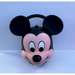 May include: A black and pink Mickey Mouse-shaped lunchbox with a handle. The lunchbox features large black ears, a pink face, white eyes, and a black nose. The lunchbox is made of plastic and has a handle on top.