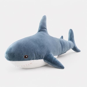 May include: A plush shark toy featuring a soft blue upper body and a white underbelly. The stuffed animal has a prominent dorsal fin, side fins, a tail, and small dark eyes. The mouth area shows a row of small, soft white teeth along the jawline.