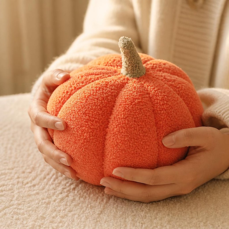 May include: A round, orange pumpkin-shaped plush toy made of textured, fuzzy fabric. The pumpkin features deep vertical segments and a small, light brown, textured stem at the top center, providing a soft, tactile appearance.