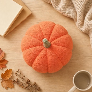 May include: A round, orange pumpkin-shaped plush pillow with a textured, fuzzy surface and a small, light green stem at the center. The pumpkin features distinct, segmented ridges radiating from the top stem down to the base of the object.