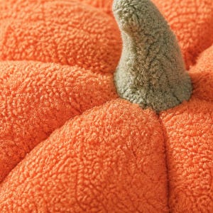 May include: A plush pumpkin-shaped object with a bright orange, textured, fleece-like fabric surface. The pumpkin features deep, rounded segments and a prominent, curved stem at the top, which is covered in a soft, muted sage green fabric.