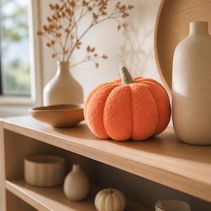 May include: A plush, textured orange pumpkin decoration with a small, light green stem. The pumpkin has a soft, fuzzy surface and is rounded with distinct vertical segments, sitting on a wooden shelf alongside various neutral-toned ceramic vases.