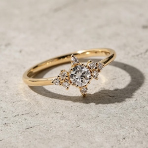 May include: A gold ring featuring a central round, clear gemstone surrounded by smaller, clear gemstones in a starburst pattern. The ring band is thin and gold. The ring is set on a light grey surface.