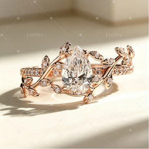 Könnte beinhalten: Ein Ring aus Roségold mit einem zentralen, birnenförmigen, klaren Edelstein. Die Ringschiene ist mit filigranen Metallblattmotiven gestaltet, die sich entlang der Seiten erstrecken und mit kleinen, runden, klaren Steinen besetzt sind. Das Wort Lunov ist sichtbar.