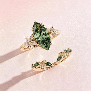 May include: A gold ring set featuring a large marquise-cut green gemstone as the centerpiece, flanked by small clear stones arranged in floral patterns. A matching curved band with leaf-shaped green stones and clear accents completes the set.