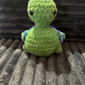 May include: A handmade, green crochet turtle with black eyes and blue accents. The plush turtle is made of a soft, textured yarn and is sitting on a gray, textured surface. The turtle is a cute, whimsical design.