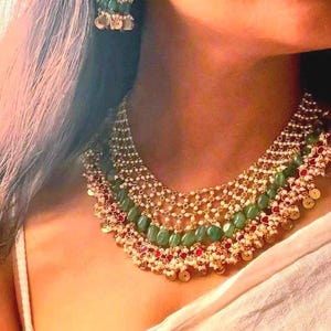 Op de afbeelding: Een meerlaagse statement ketting met rijen kleine witte parels, ovaalvormige groene kralen en kleine rode accenten. Het ontwerp bevat ingewikkeld goudkleurig metaalwerk met bungelende ronde bedels langs de onderste rand.