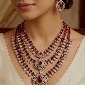 Op de afbeelding: Een driedelige ketting en bijpassende oorbellenset. De sieraden hebben een combinatie van rode edelstenen en heldere, sprankelende accenten. De ketting heeft een gelaagd ontwerp met sierlijke hangers. De oorbellen weerspiegelen het ontwerp van de ketting.