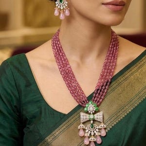 May include: A multi-strand necklace featuring pink oval beads and a central pendant with green stones, clear crystals, and dangling pink teardrop beads. Matching earrings feature a green stone, clear crystals, and three pink teardrop beads.