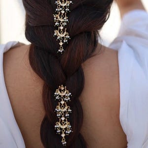May include: Two gold-toned hair accessories featuring a vertical arrangement of crescent moon shapes, clear stones, and small white beads. Each piece is designed with a series of dangling elements that hang down the length of a braided hairstyle.