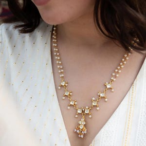May include: A gold necklace with a detailed design. The necklace has small, square-shaped features with clear stones and tiny pearls. A cluster of pearls and stones hangs from the centre, creating an elegant look. The necklace is worn against a white top.