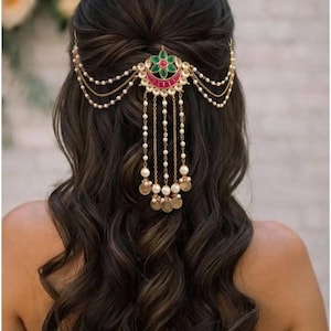 May include: An ornate gold-toned hair accessory with a central design of green and red gemstones, accented by pearls and dangling coin-shaped charms. The headpiece is set against dark, wavy hair, suggesting a bridal or formal occasion.