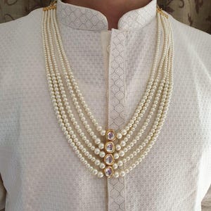 Peut inclure: Un collier de perles à plusieurs rangs avec des détails dorés et trois pierres claires de forme ovale. Le collier est porté sur une chemise blanche texturée avec un col officier. Les perles sont de couleur blanc crème.
