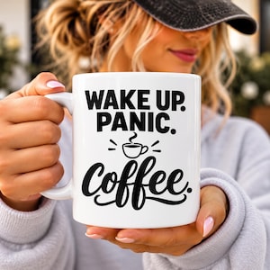 May include: A white ceramic mug featuring the black printed text WAKE UP. PANIC. followed by a small coffee cup icon and the word coffee in a cursive script font. The mug has a standard handle and a smooth, glossy finish.