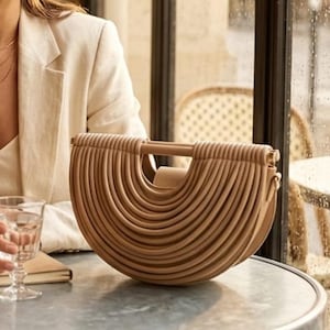 May include: A beige, semi-circular handbag featuring a structured, ribbed exterior design with a horizontal top handle. The bag has a curved, layered appearance with a solid, smooth finish, presented in a neutral tone with metallic hardware accents.