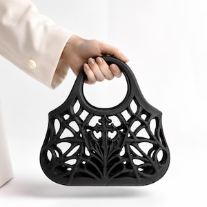 May include: A matte black handbag featuring a circular handle and an intricate, openwork lattice design that resembles a spider web. The centre of the bag displays a prominent, raised cross motif integrated into the structural web pattern.