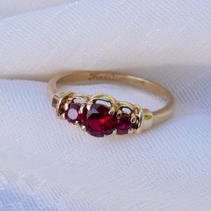 Könnte beinhalten: Ein Goldring mit drei runden, tiefroten facettierten Steinen in einer Krappenfassung. Der Ring ist glatt und poliert, wobei das Wort Tradition und die Kennzeichnung 10K auf der Innenseite des Goldrings eingraviert und im Licht sichtbar sind.