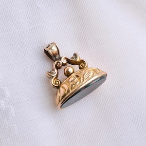 May include: A gold-toned metal wax seal pendant featuring an ornate, scrolled handle with a central sphere. The base of the seal is circular and decorated with intricate engraved floral patterns, holding a smooth, dark, reflective stone insert.