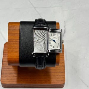 May include: A rectangular silver-tone wristwatch with a black leather strap featuring a textured sunburst pattern on the dial and case. The white face displays black Arabic numerals, the text IBSO, and blue hands, with a crown on the side.