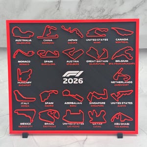 May include: A rectangular black display board with a red frame featuring raised red outlines of twenty-four Formula 1 race tracks. Each track is labeled with its country and city name in white text. The center displays the white text F1 2026.
