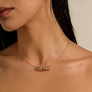 以下が含まれることがあります： 「EMMA」という名前が個別の文字で書かれたゴールドカラーのネックレス。ネックレスは首の周りに着用され、チェーンと文字が見えます。文字はブロック体です。