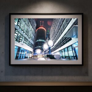 Könnte beinhalten: Gerahmter Druck einer modernen Stadtlandschaft bei Nacht. Das Bild zeigt ein hohes, zylindrisches Gebäude mit roter Spitze, flankiert von moderner Architektur. Straßenlaternen beleuchten den Vordergrund, und der Himmel ist dunkel mit Bokeh.