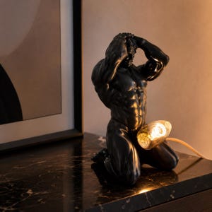 May include: Black sculpture of a kneeling male figure with arms raised, holding his head. A light bulb is positioned in front of the figure. The sculpture is on a dark marble surface.