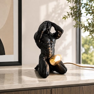 May include: Black sculpture of a muscular male figure kneeling with arms raised. A light bulb is positioned in the groin area, emitting a warm glow. The lamp is on a marble surface.