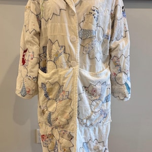May include: Cream-colored quilted coat with floral patchwork details. The coat features a notched collar, two front pockets, and large white buttons. The patchwork includes blue, red, and patterned fabrics, with black stitching outlining the floral shapes.