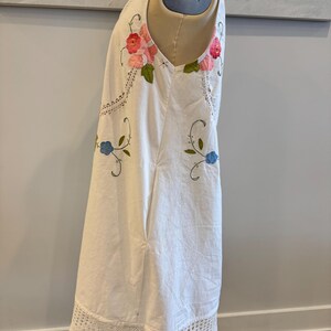 May include: White sleeveless dress with floral embroidery in pink, red, and blue. The dress features a crochet lace hem and accents. The dress is displayed on a mannequin.