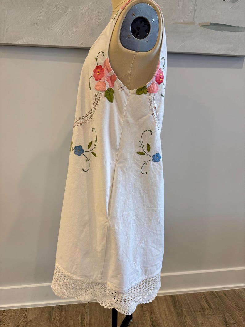 May include: White sleeveless dress with floral embroidery in pink, red, and blue. The dress features a crochet lace hem and accents. The dress is displayed on a mannequin.