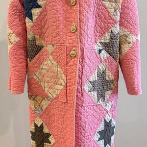 May include: A long, quilted coat in a soft pink hue, featuring a patchwork design with star and diamond patterns in various colors. The coat has a collared neckline, wooden buttons, and two front pockets. The coat is displayed on a mannequin.