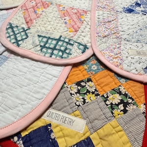 May include: Close-up of several handmade quilted pot holders. Each pot holder features a unique patchwork design with various fabric patterns and colors, including floral, plaid, and solid hues. The edges are finished with a contrasting pink or red binding. The text "QUILTED POETRY" is visible on a label.
