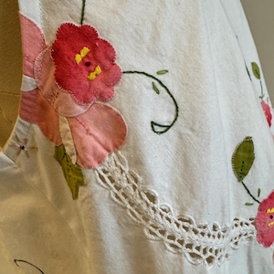 May include: Close-up of a white garment with floral embroidery. The embroidery features pink and red flowers with yellow centers, green leaves, and a white lace trim. The garment appears to be made of a textured fabric.