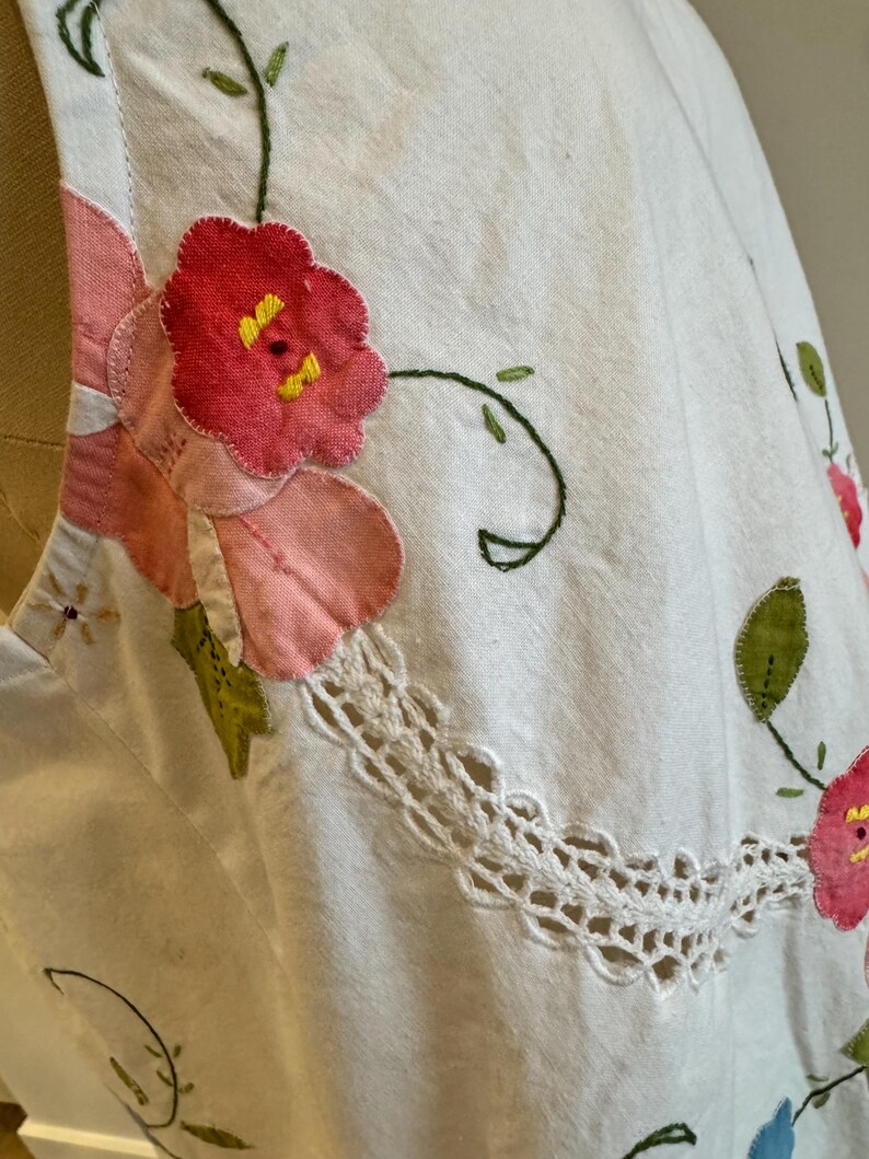 May include: Close-up of a white garment with floral embroidery. The embroidery features pink and red flowers with yellow centers, green leaves, and a white lace trim. The garment appears to be made of a textured fabric.