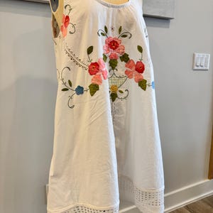 May include: White sleeveless dress with floral embroidery in pink, red, and green. The dress features a scoop neckline and a crochet lace hem. The floral design is centered on the front, with additional embroidery along the neckline.